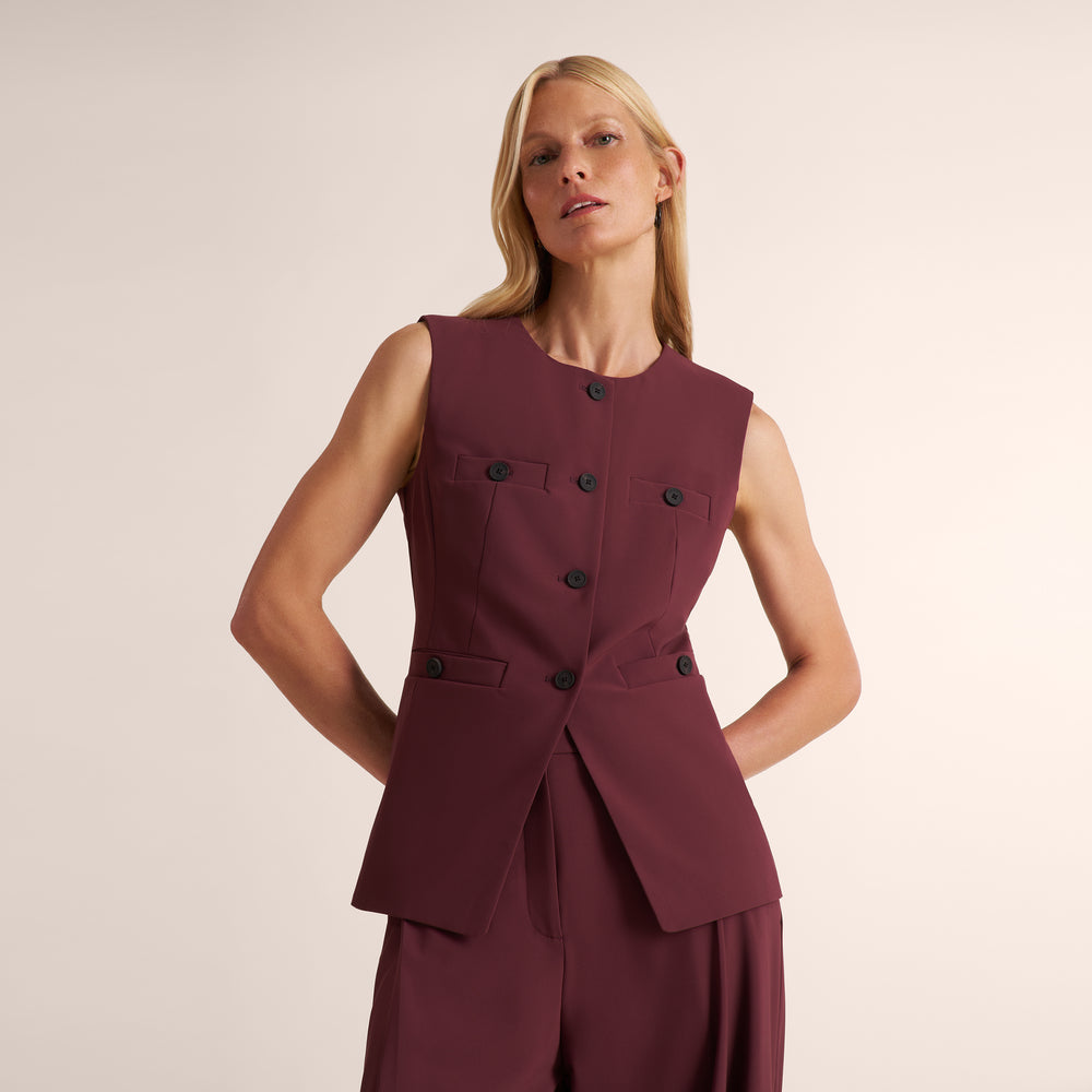 A blonde model wearing a sleeveless, structured burgundy vest with black buttons and matching trousers, posed against a solid light beige studio background.