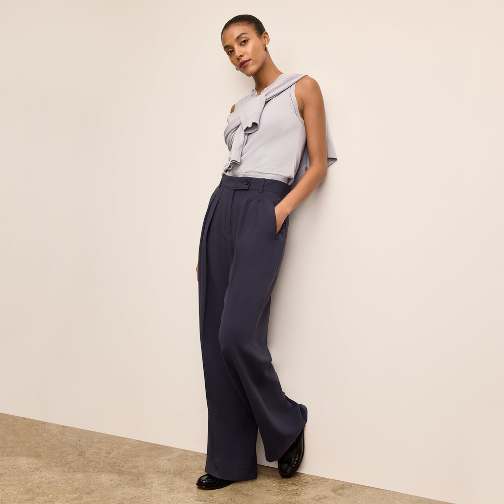 Woman wearing a light gray sleeveless top and dark blue high-waisted pants against a beige wall.