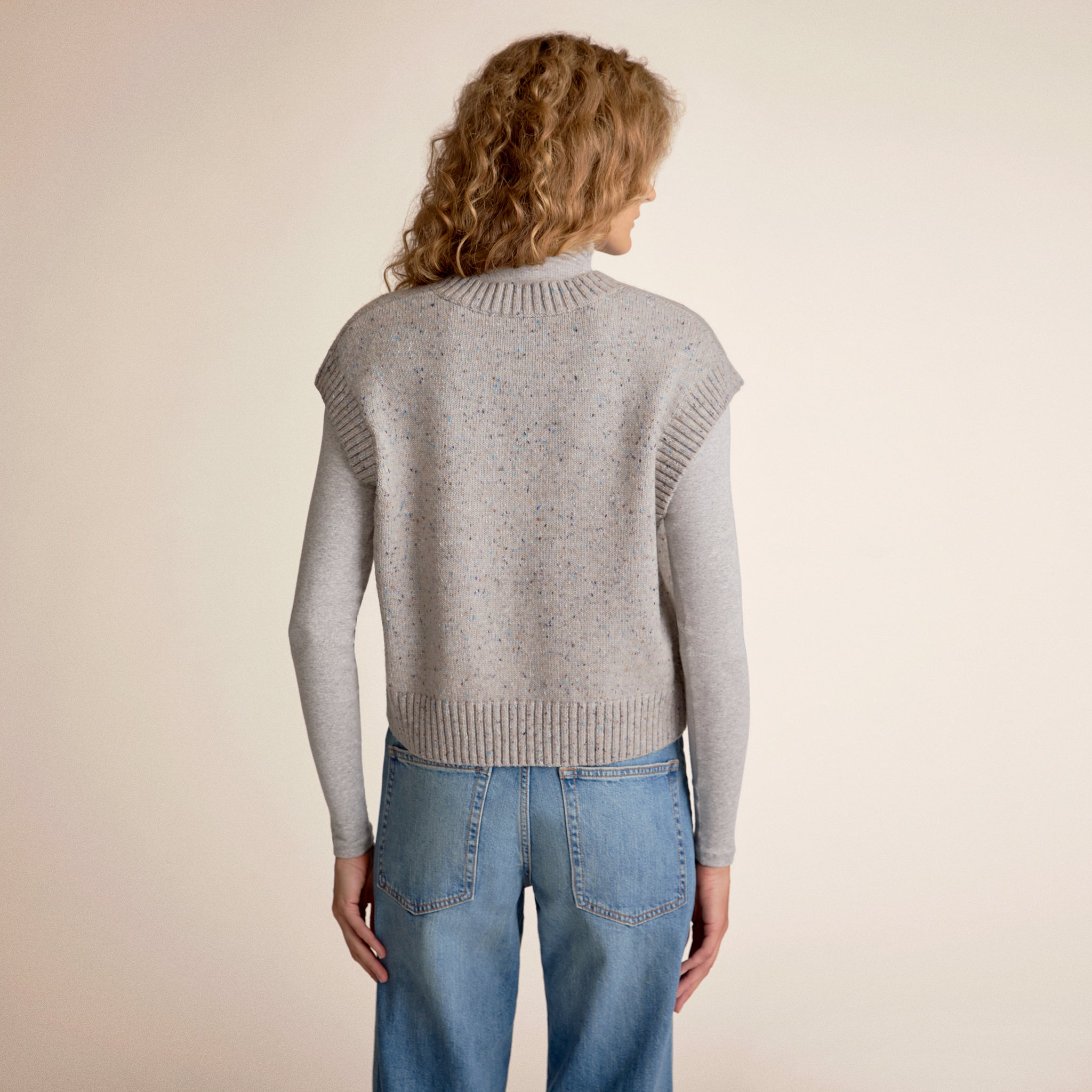 Person wearing a gray sweater vest and blue jeans on a beige background