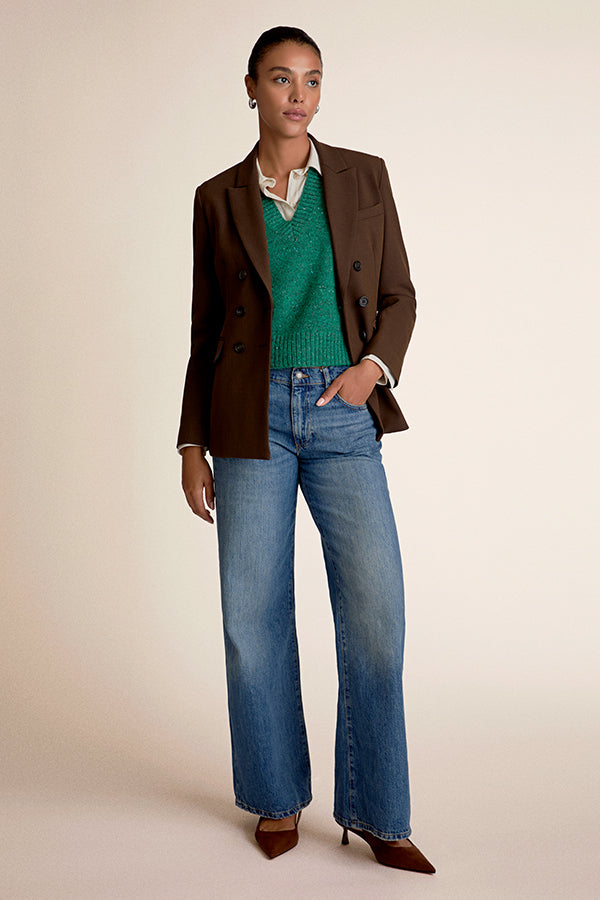 Woman wearing a brown jacket, green sweater, and blue jeans on a beige background