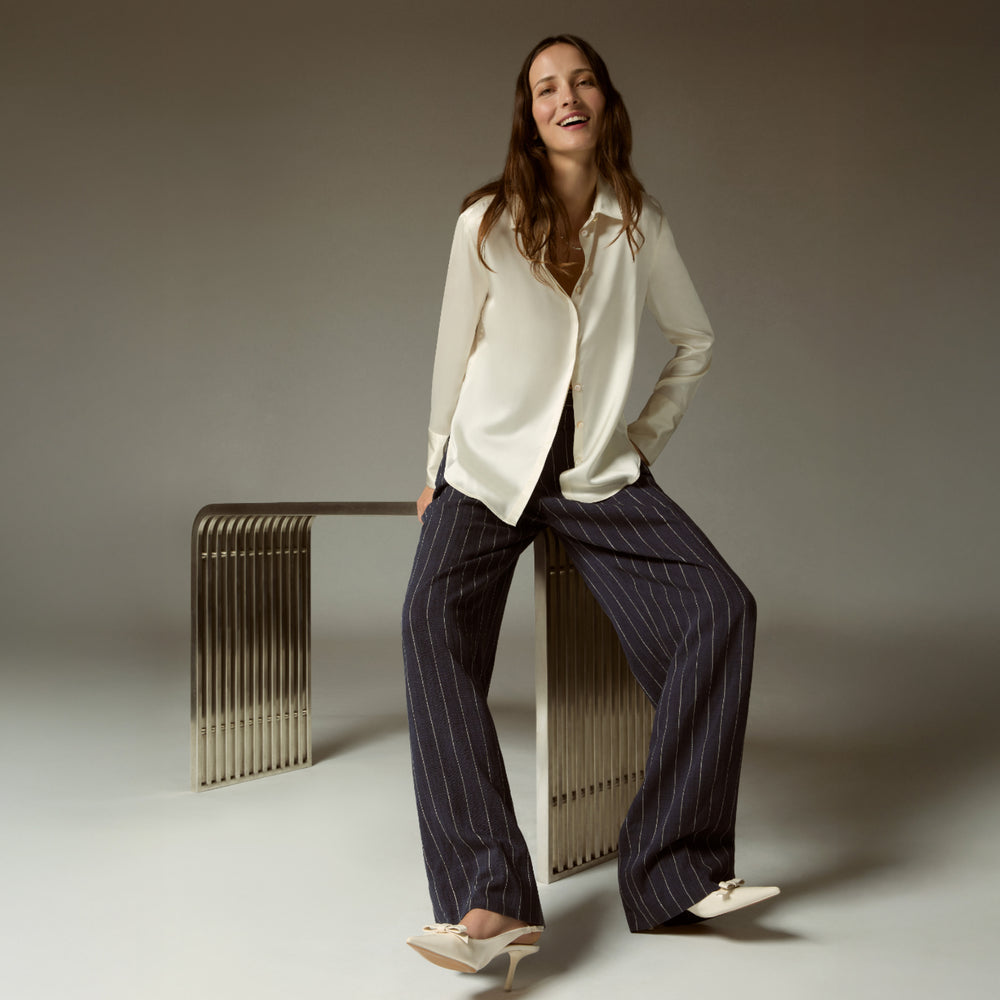 Woman in a white shirt and striped pants sitting on a metallic bench against a neutral background