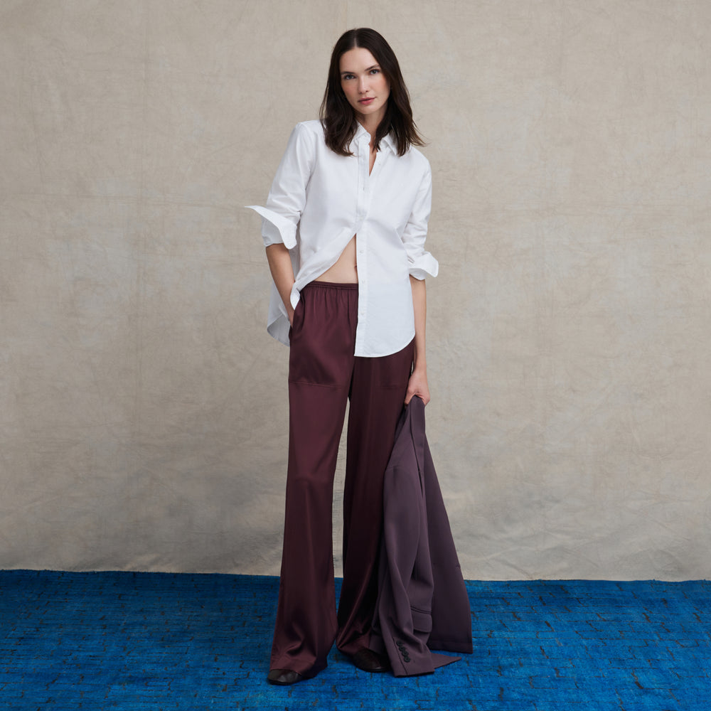 Woman wearing a long sleeve white button down with silk aubergine wide leg pants holding a purple blazer on a blue carpet floor and against a beige background