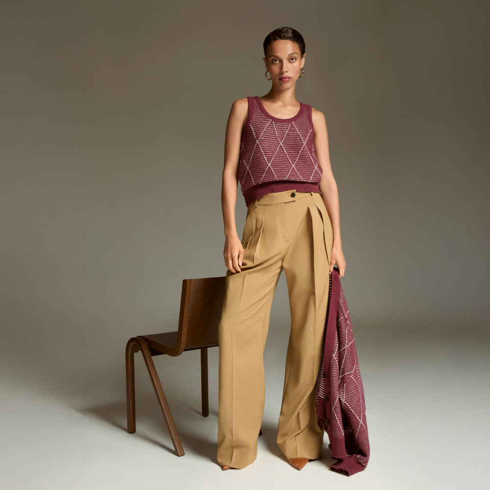 Woman wearing a patterned sleeveless top and beige pants, holding a matching fabric, against a neutral background.