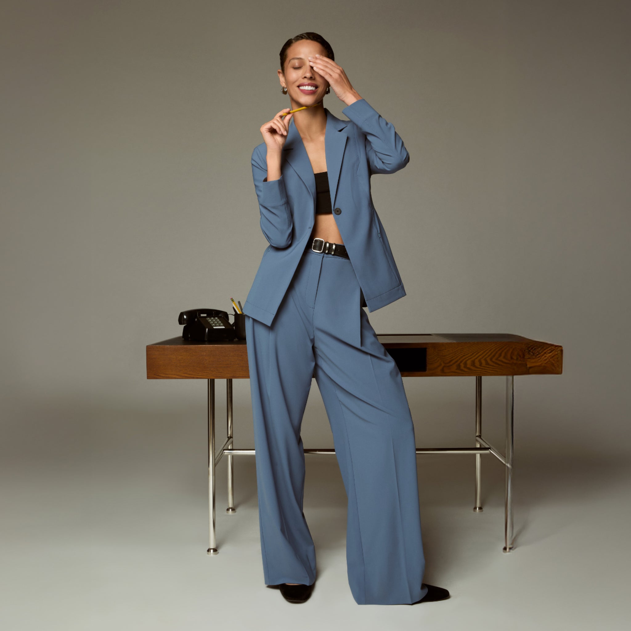 Woman in a blue suit standing next to a wooden desk with a phone on it.