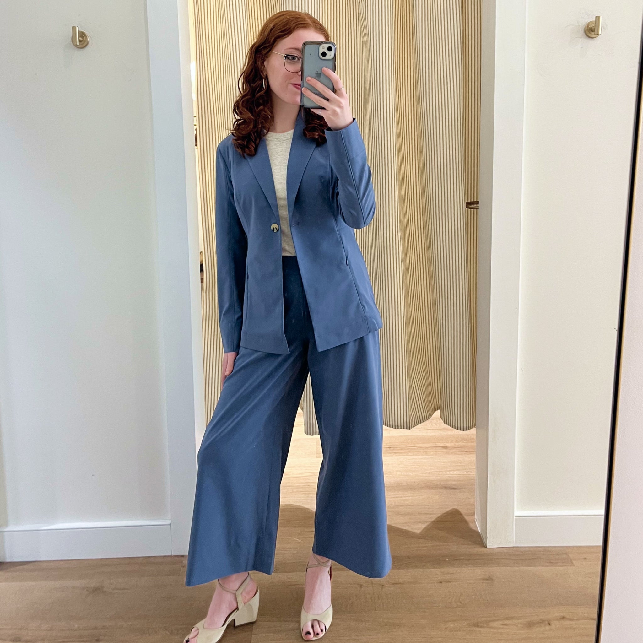 Person wearing a blue suit taking a mirror selfie in a room with wooden flooring and white walls.