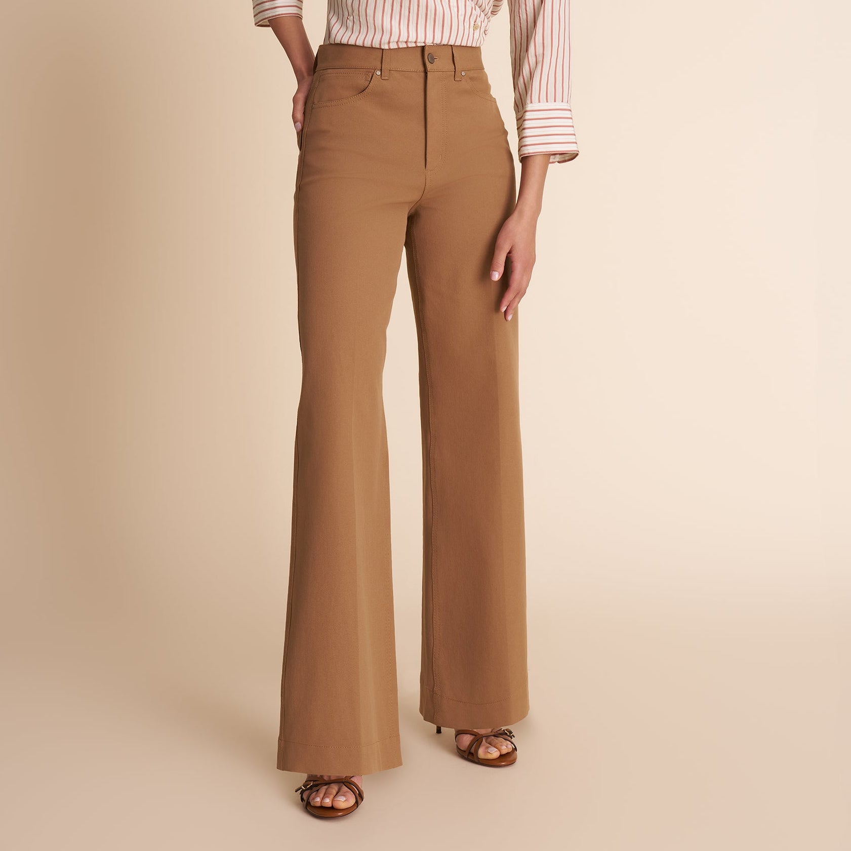 A model stands against a solid beige studio background wearing a white button-down shirt with thin red vertical stripes, tucked into high-waisted, wide-leg brown trousers, and brown sandals.