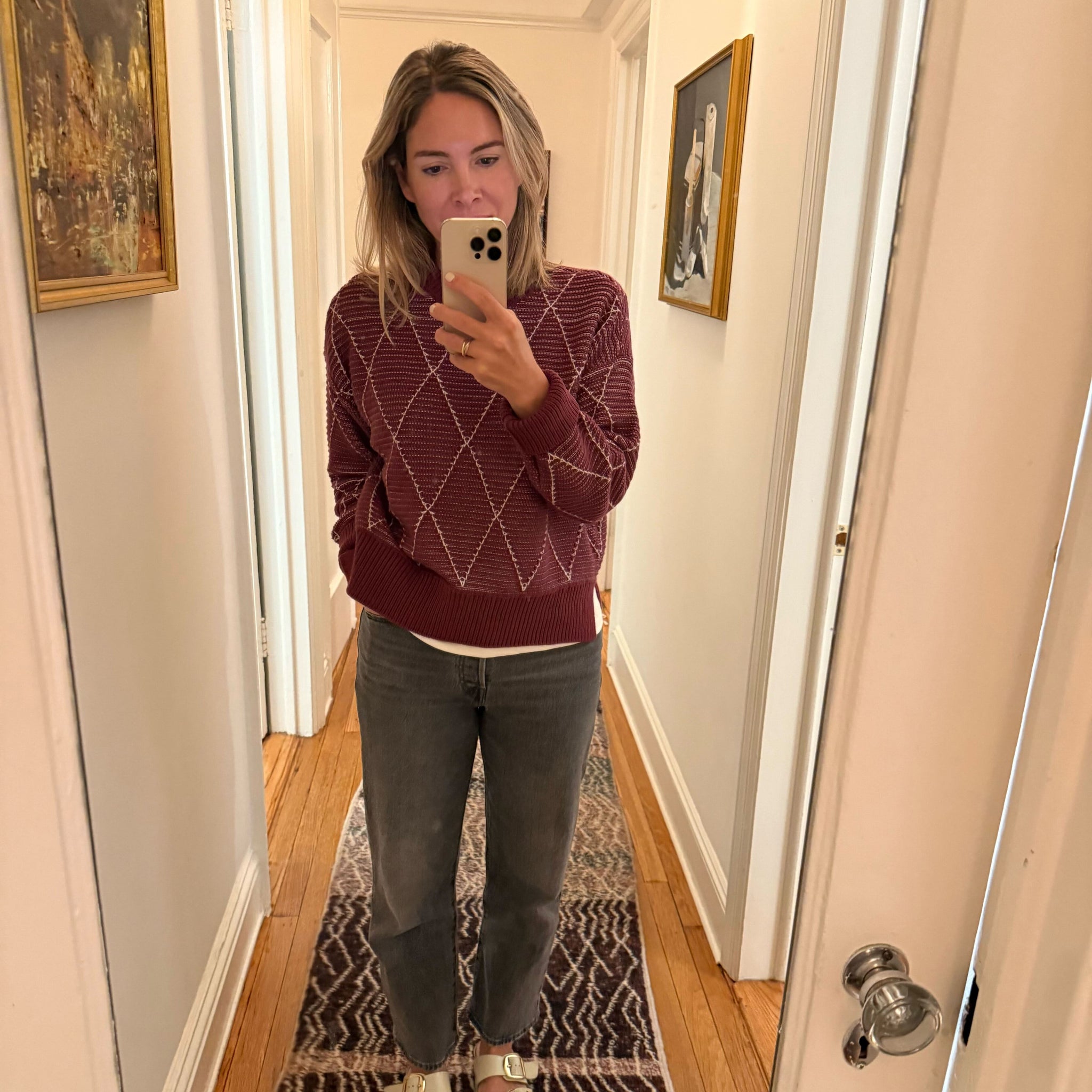 Person taking a mirror selfie in a hallway wearing a maroon sweater and gray pants.