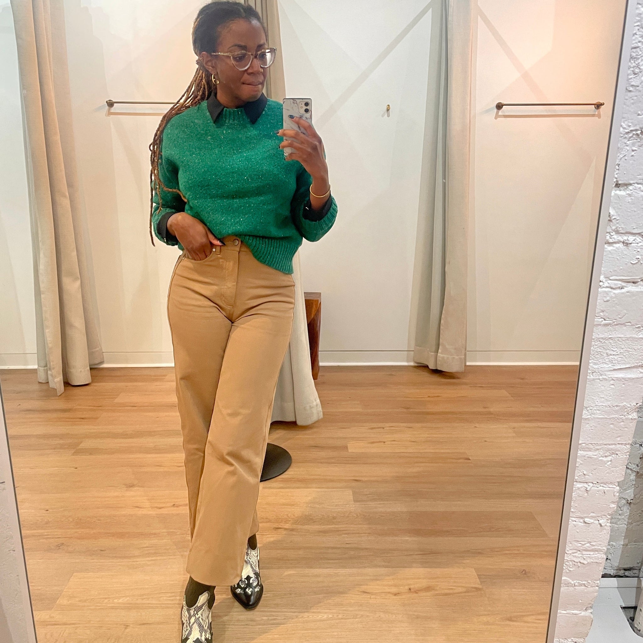 Person taking a mirror selfie wearing a green sweater and beige pants in a room with wooden flooring.