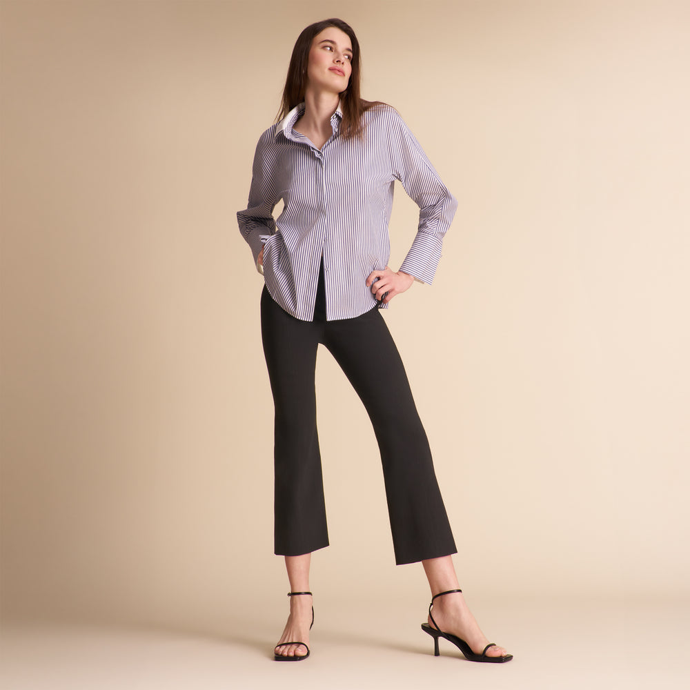 A woman models a light purple and white striped button-down shirt paired with black cropped pants and black strappy heels, set against a solid beige studio background.