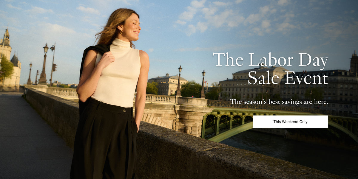 A woman in a cream sleeveless turtleneck and black skirt stands on a stone bridge in Paris with the Seine river and historic buildings in the background. Large promotional text on the right reads 'The Labor Day Sale Event' with a button below.