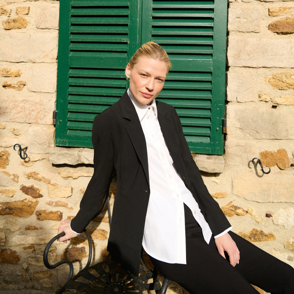 Woman in black coat and white shirt sitting on a chair against a stone wall with green shutters.