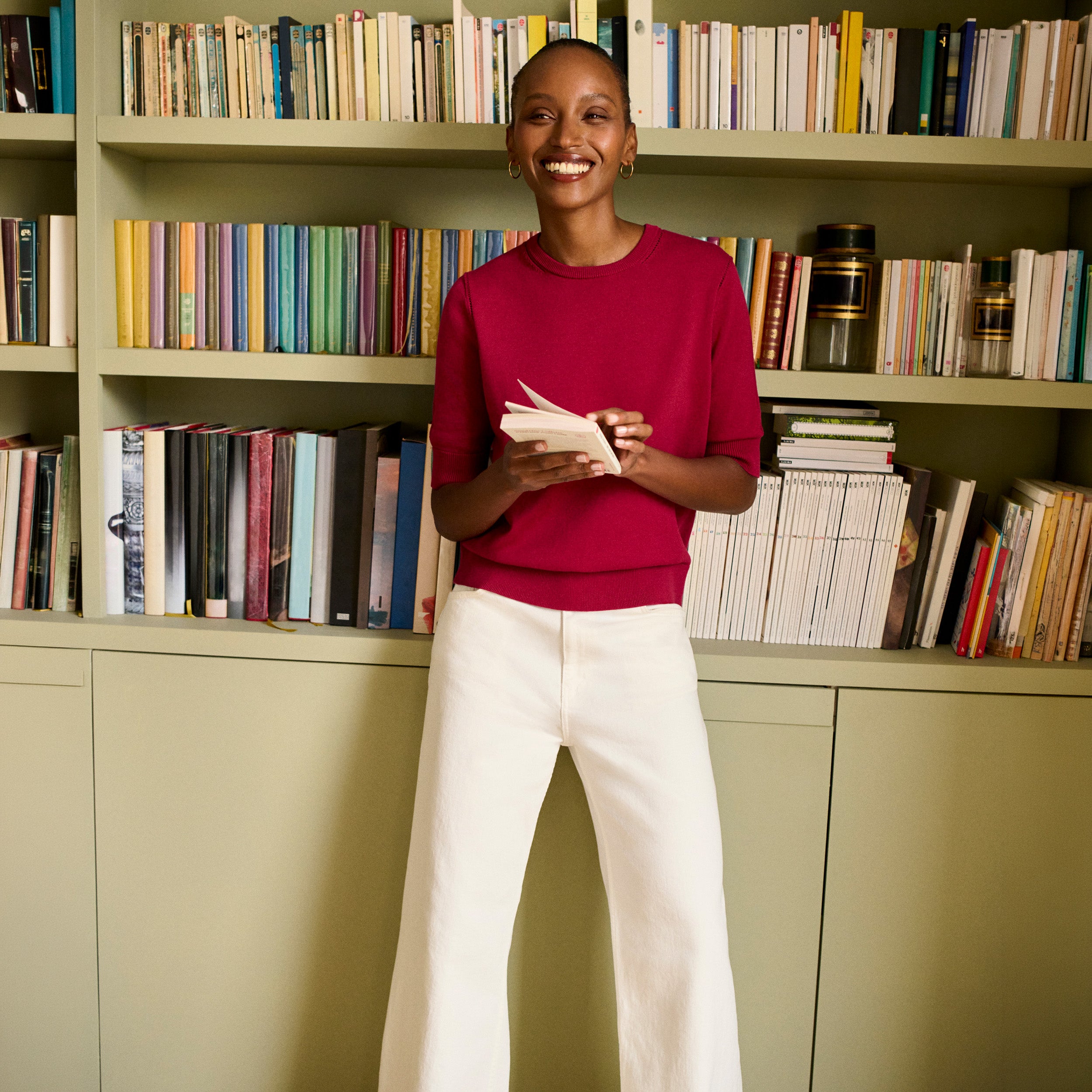 A woman stands in front of a bookshelf wearing a bright pinkish-red long-sleeve top and white wide-leg pants, holding a small book or notebook.