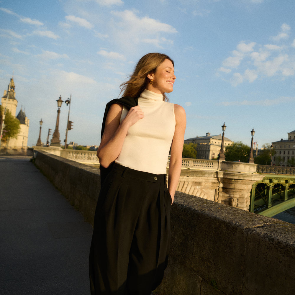 Woman in a cream top and black pants standing on a bridge with a cityscape background
