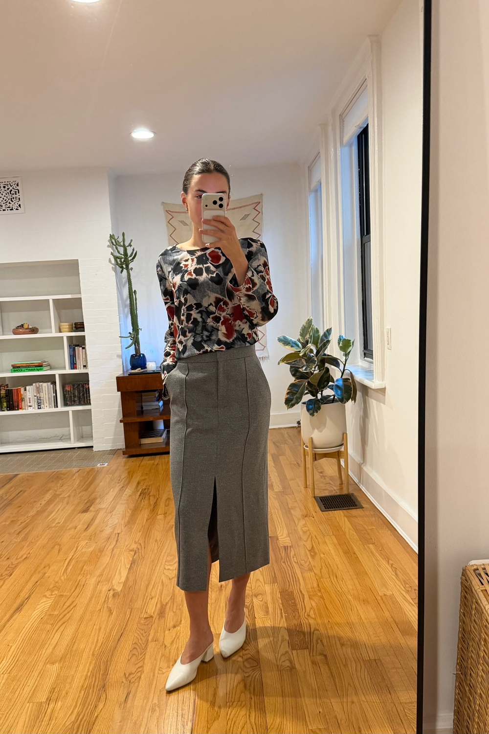 Person taking a mirror selfie wearing a floral top and gray skirt in a room with wooden flooring.