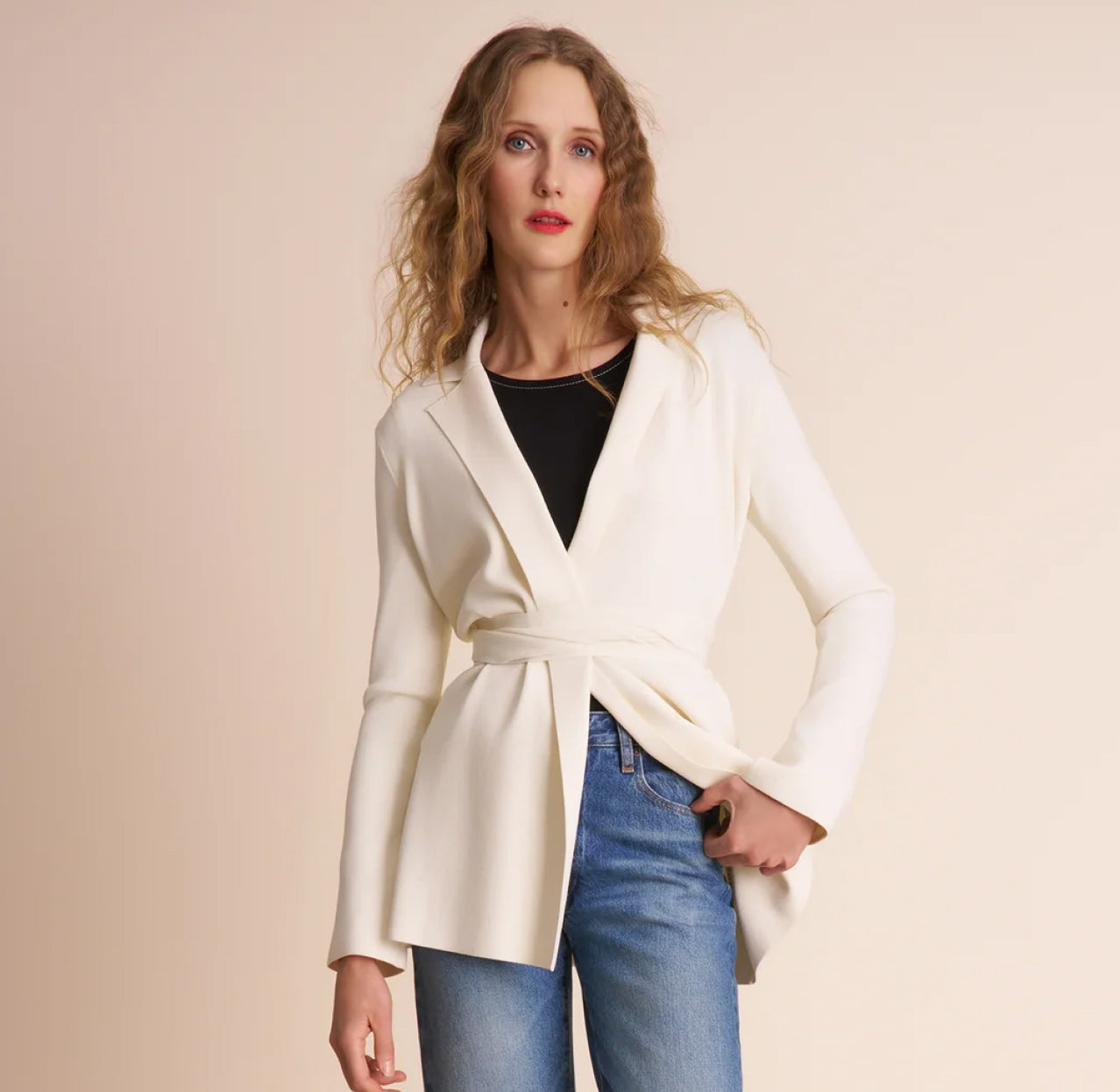Woman wearing a white wrap coat over a black top and blue jeans on a beige background