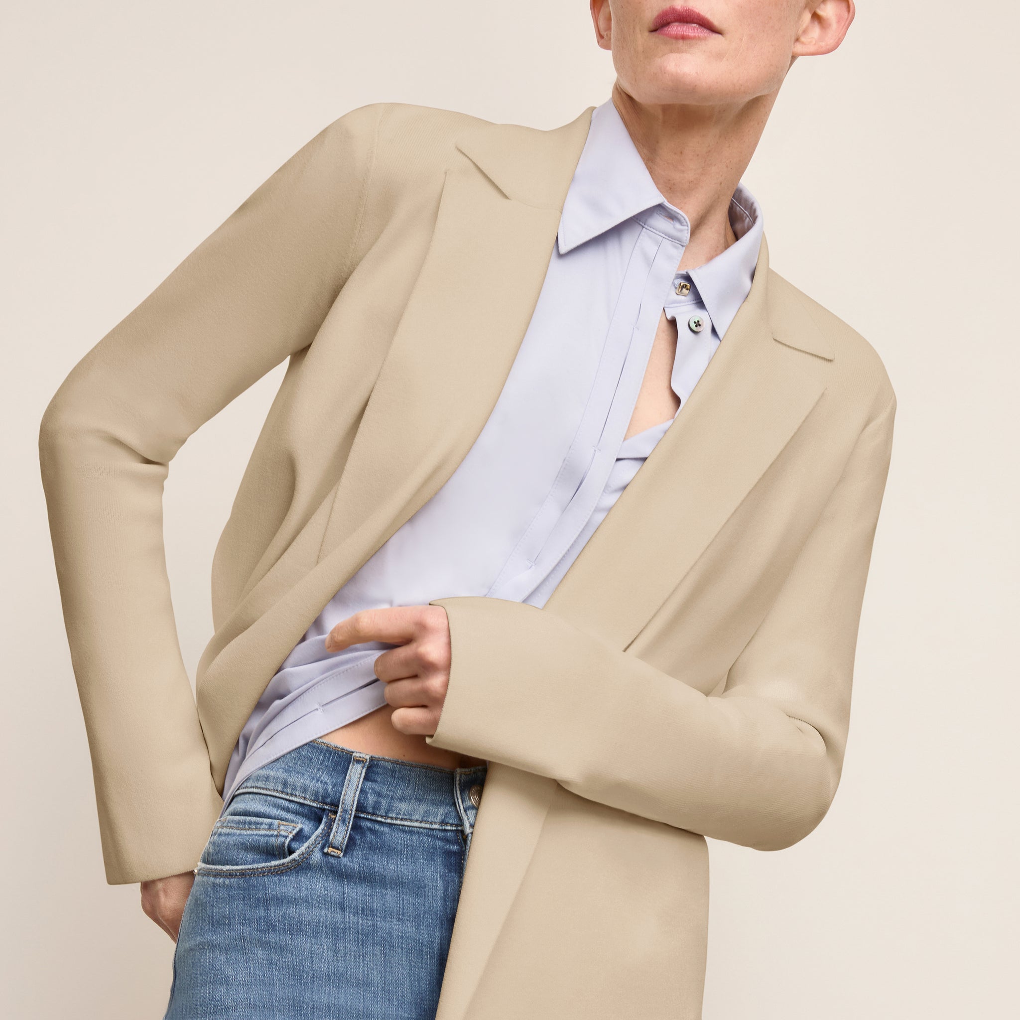 Person wearing a beige blazer over a light blue shirt and jeans on a beige background