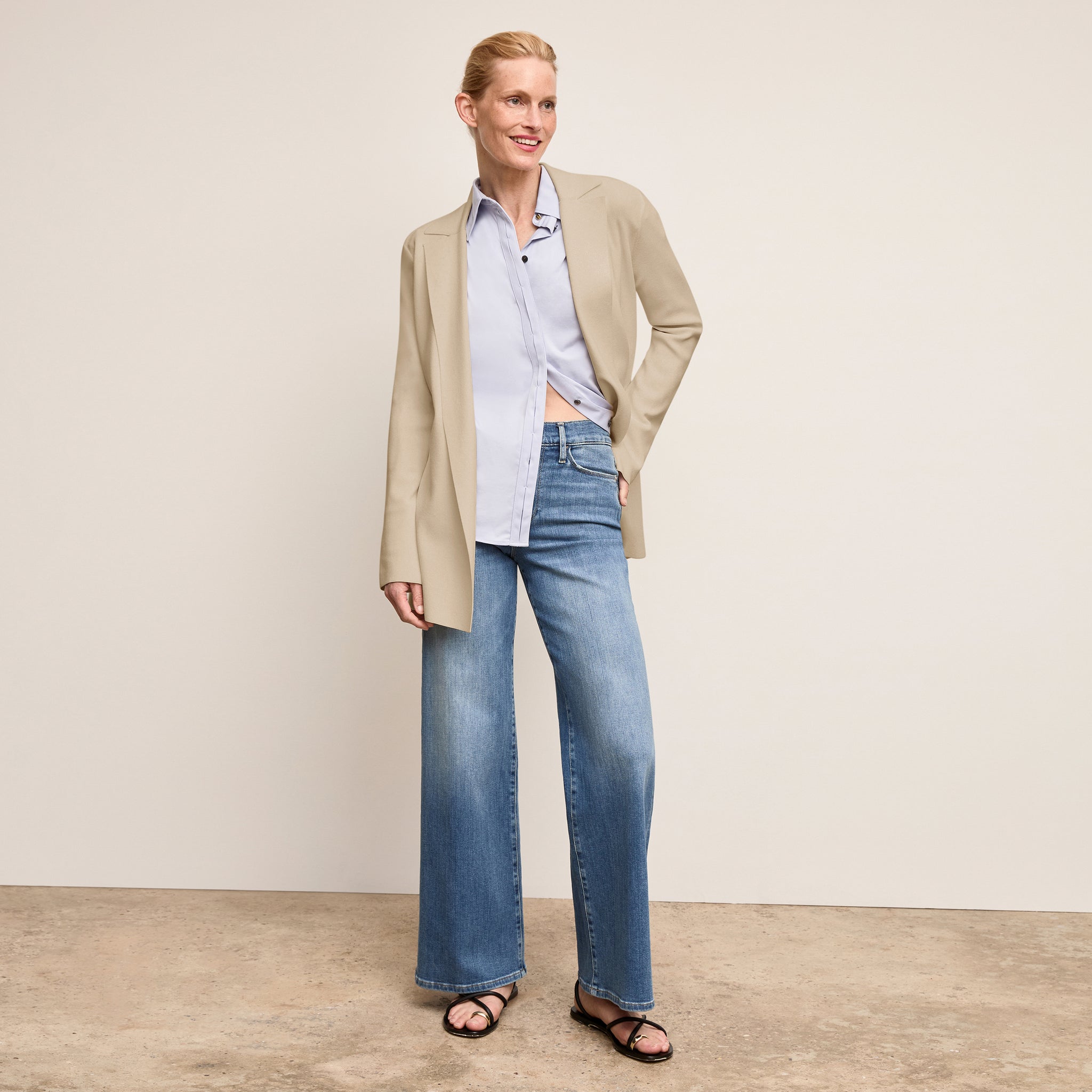 Woman wearing a beige cardigan, light blue shirt, and jeans against a plain background