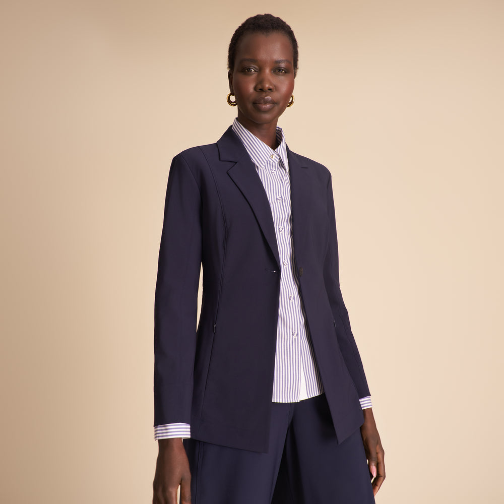 Woman in navy blazer with matching pants and striped shirt sitting against a beige background