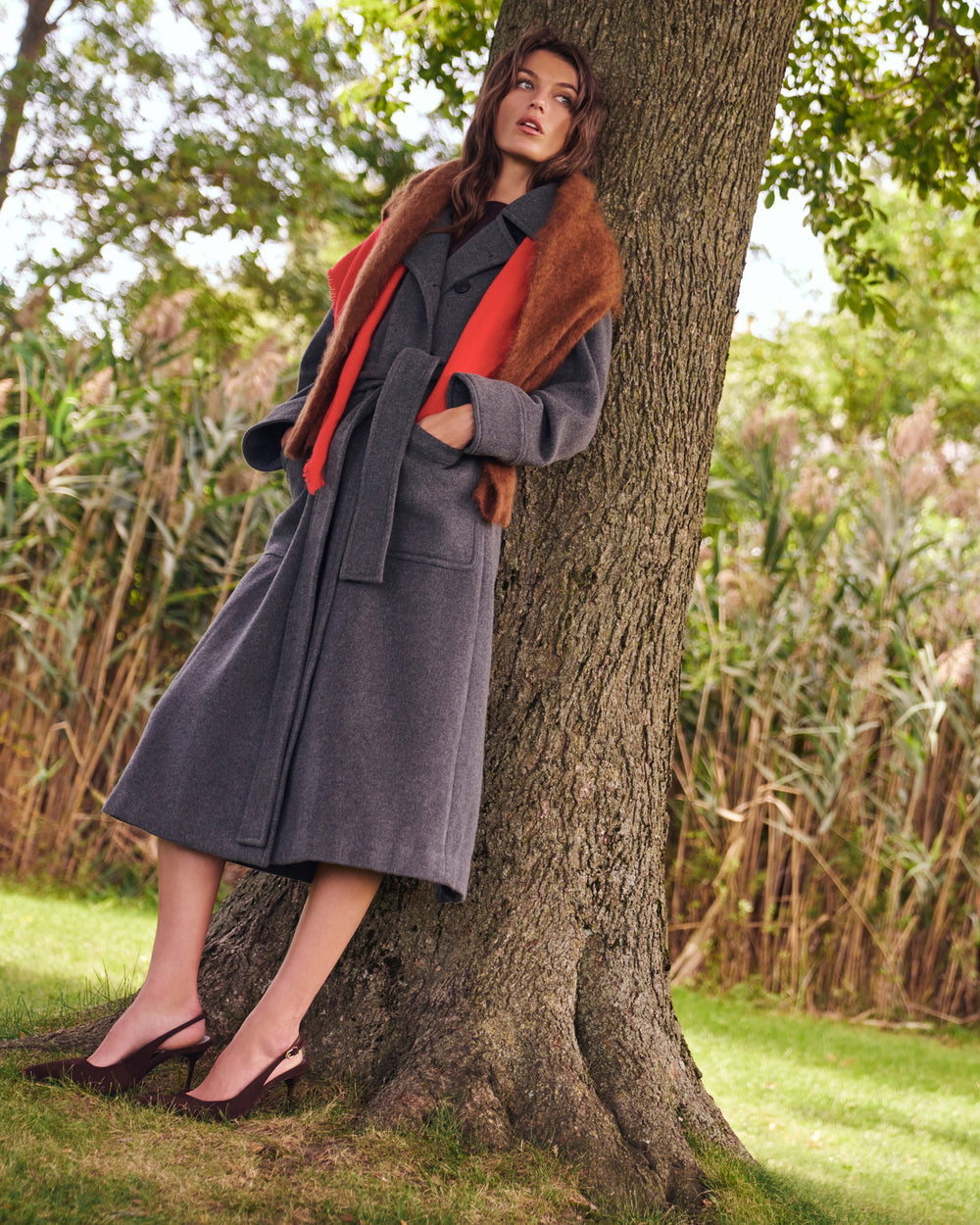 A woman leans against a large tree outdoors, wearing a long charcoal gray coat with a brown fur-trimmed collar and a bright orange-red inner layer or lining. She is wearing dark brown slingback heels, with grass, reeds, and trees in the background.