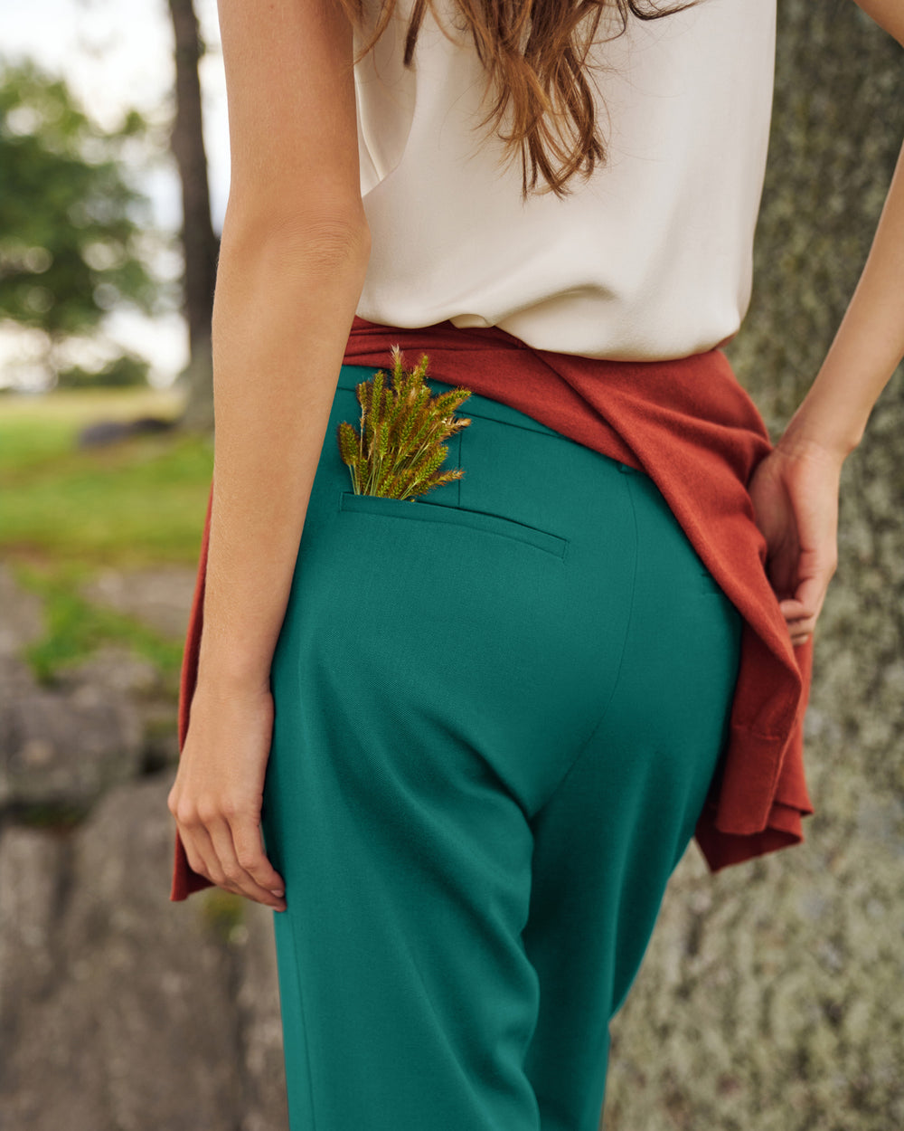A person outdoors wearing teal green trousers with a rust red cardigan draped over the waist and a cream top. The photo is cropped from the back, showing the side pocket holding a small green plant.