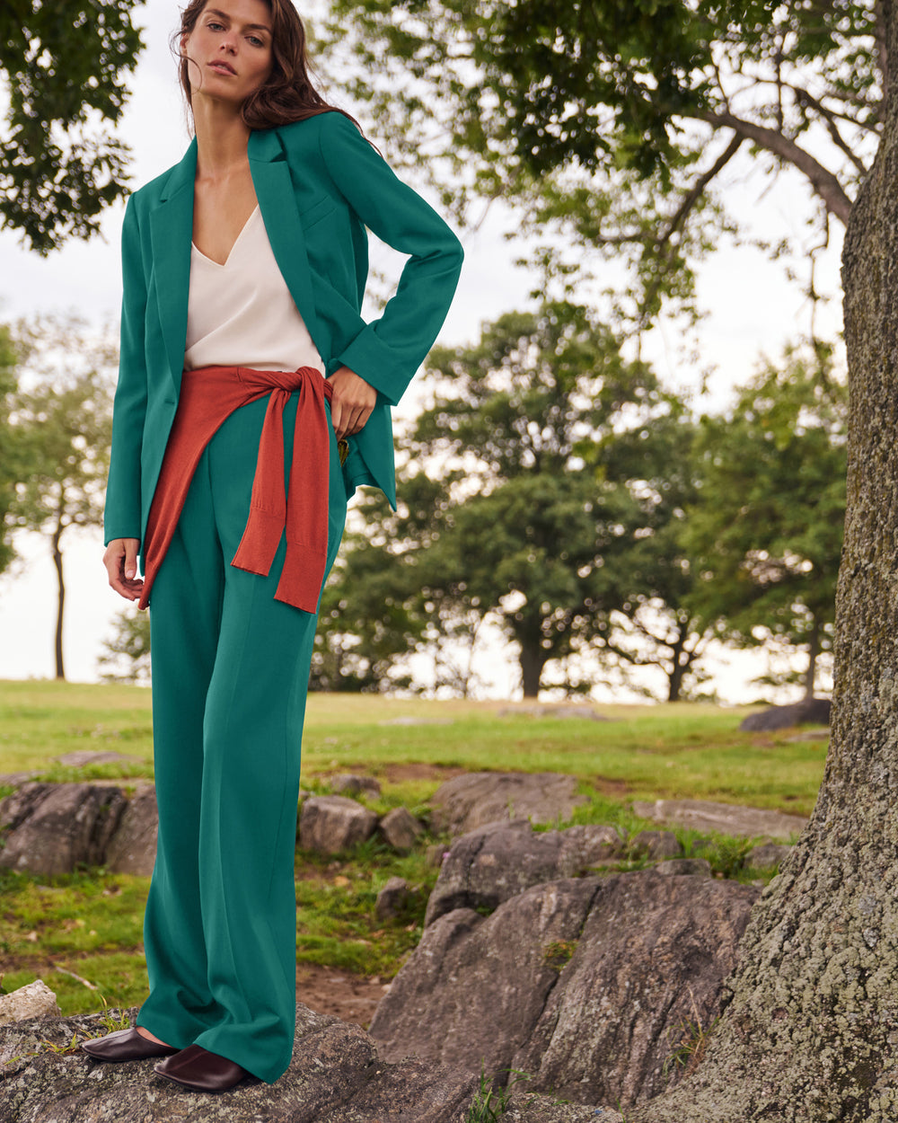 A woman stands outdoors on rocks near a tree wearing a teal green tailored blazer and matching wide-leg trousers, styled with a white top and a rust-red tied sash. The background shows a softly blurred park setting with grass, trees, and large stones.