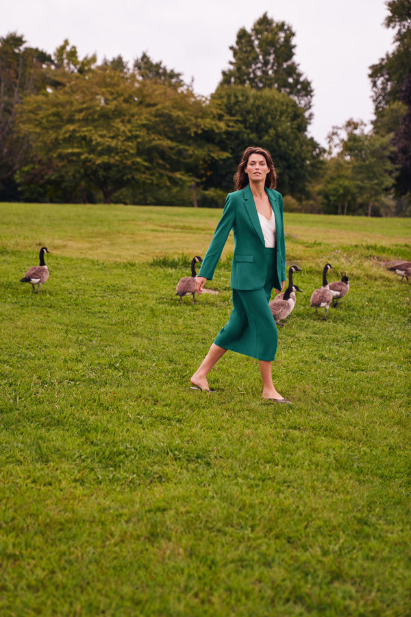 A woman in a matching emerald green two-piece skirt suit walks across a grassy field with several Canada geese in the background.