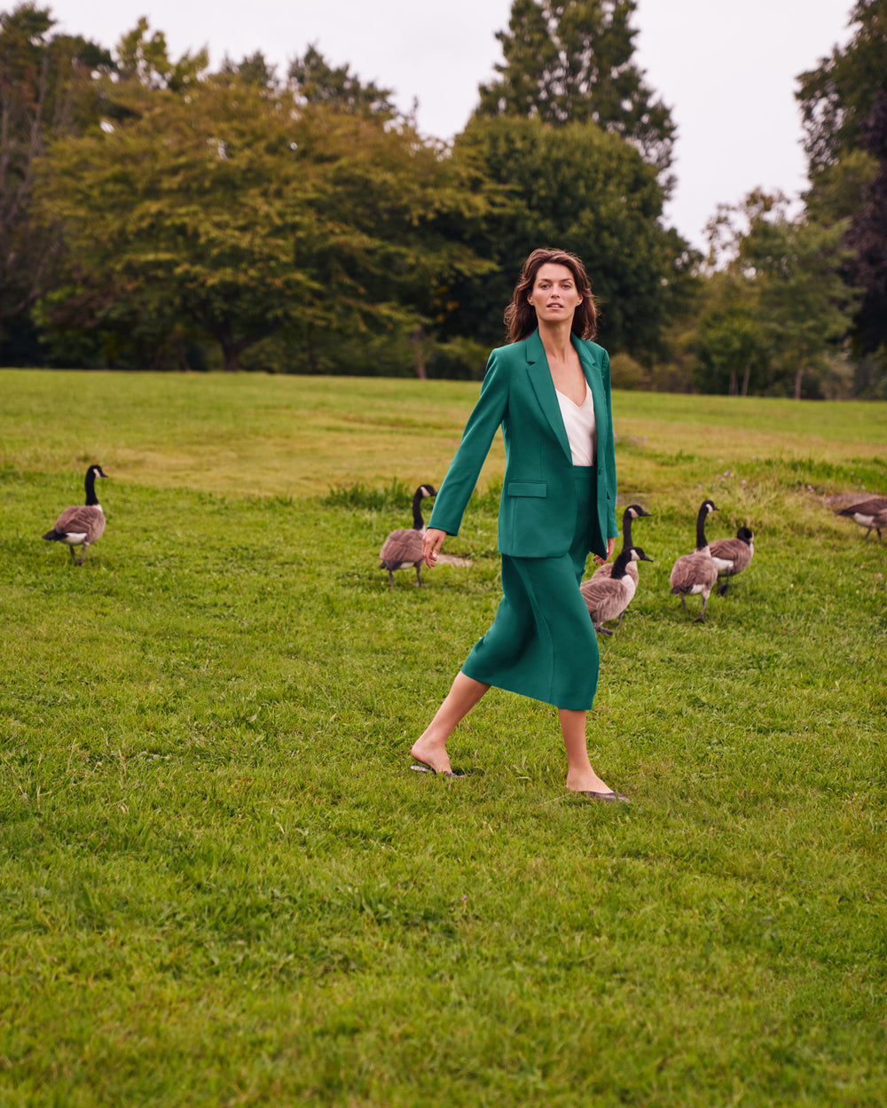 A woman walks barefoot across a grassy field wearing a matching emerald green two-piece skirt suit over a white top, with several geese standing in the background near trees.