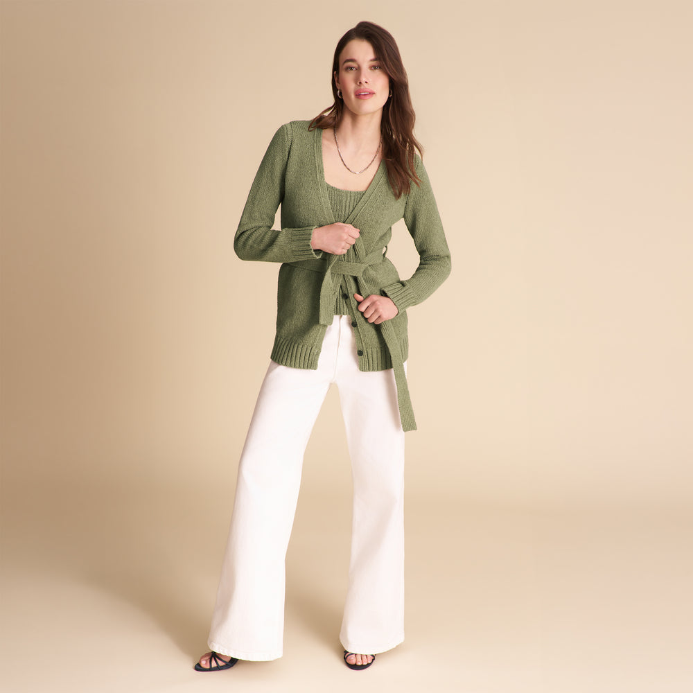 Woman wearing a green cardigan and white pants on a beige background