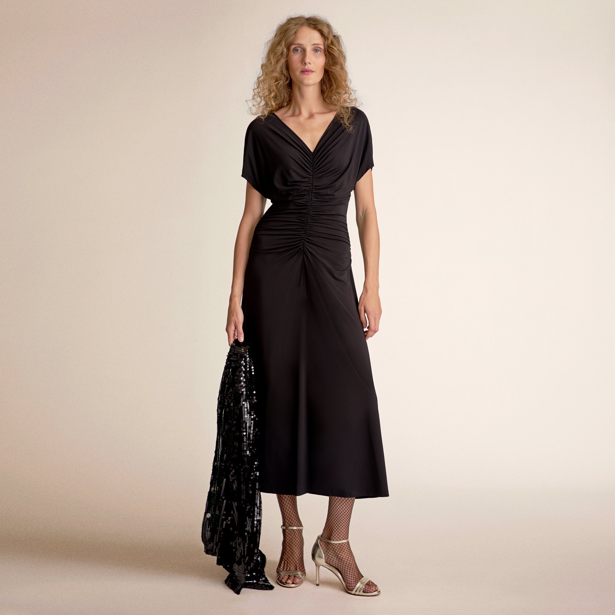 Woman wearing a black dress holding a black sequined jacket against a beige background 
