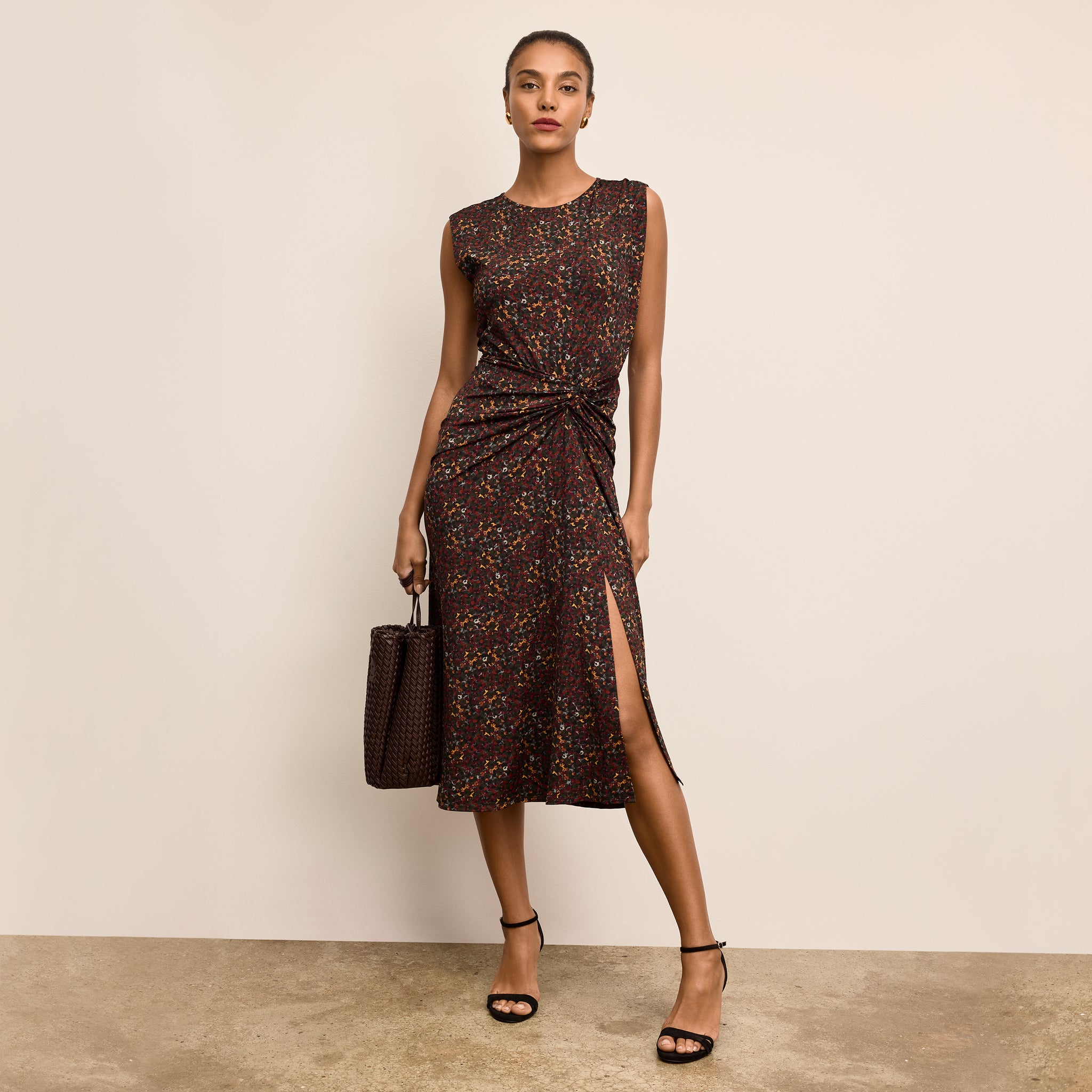 Woman wearing a patterned dress with a slit, holding a matching handbag against a beige background. 
