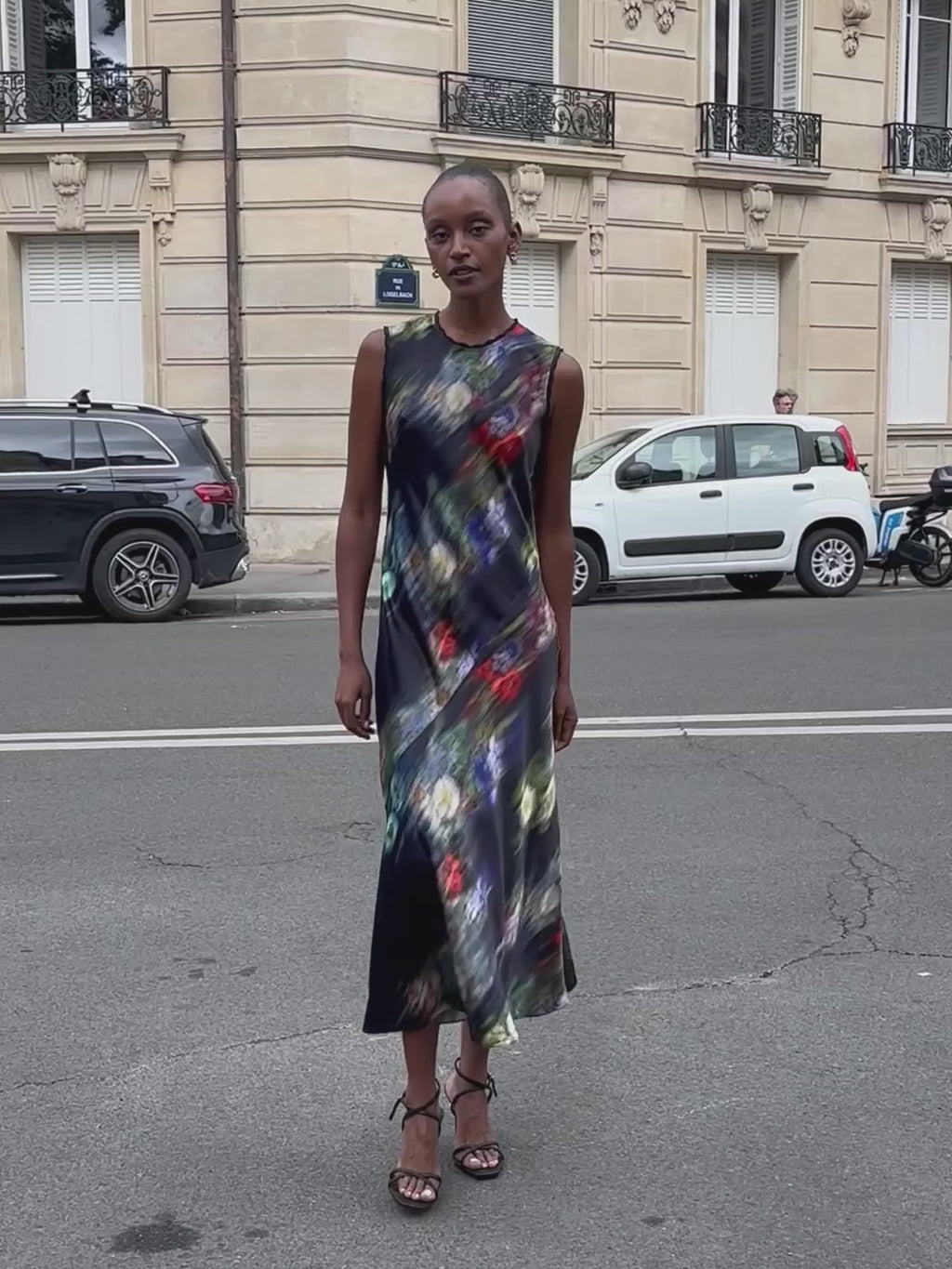 A woman modeling a sleeveless maxi dress with a dark navy base and a vibrant, multicolored abstract floral print on the city streets of Paris