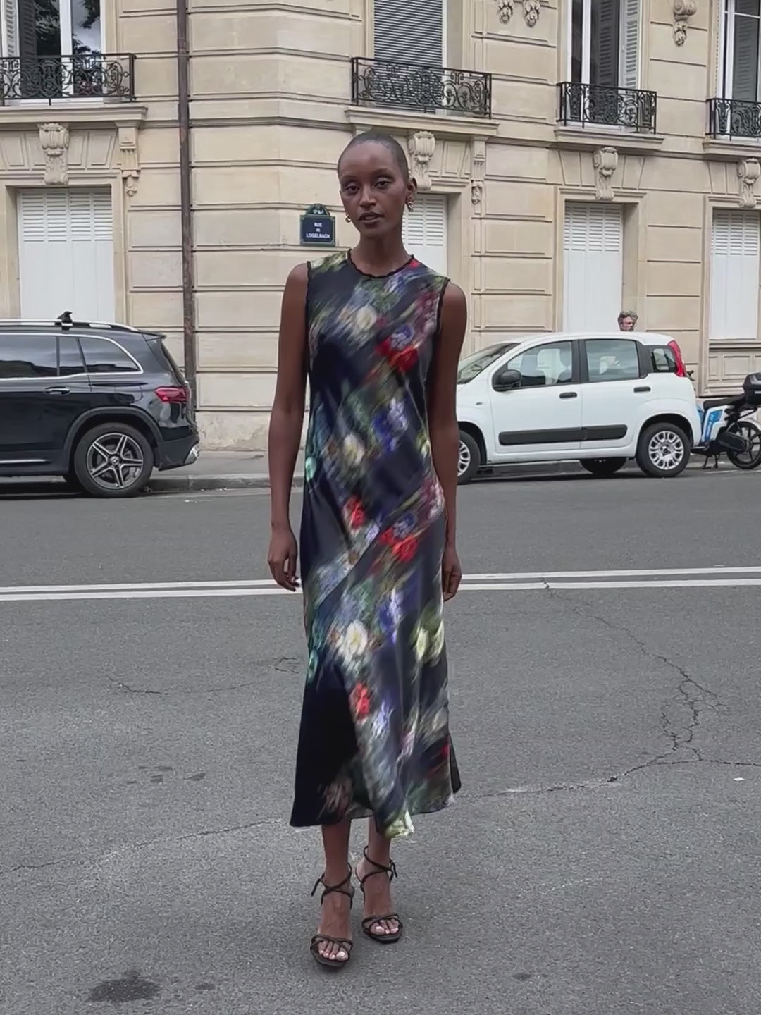 A woman modeling a sleeveless maxi dress with a dark navy base and a vibrant, multicolored abstract floral print on the city streets of Paris